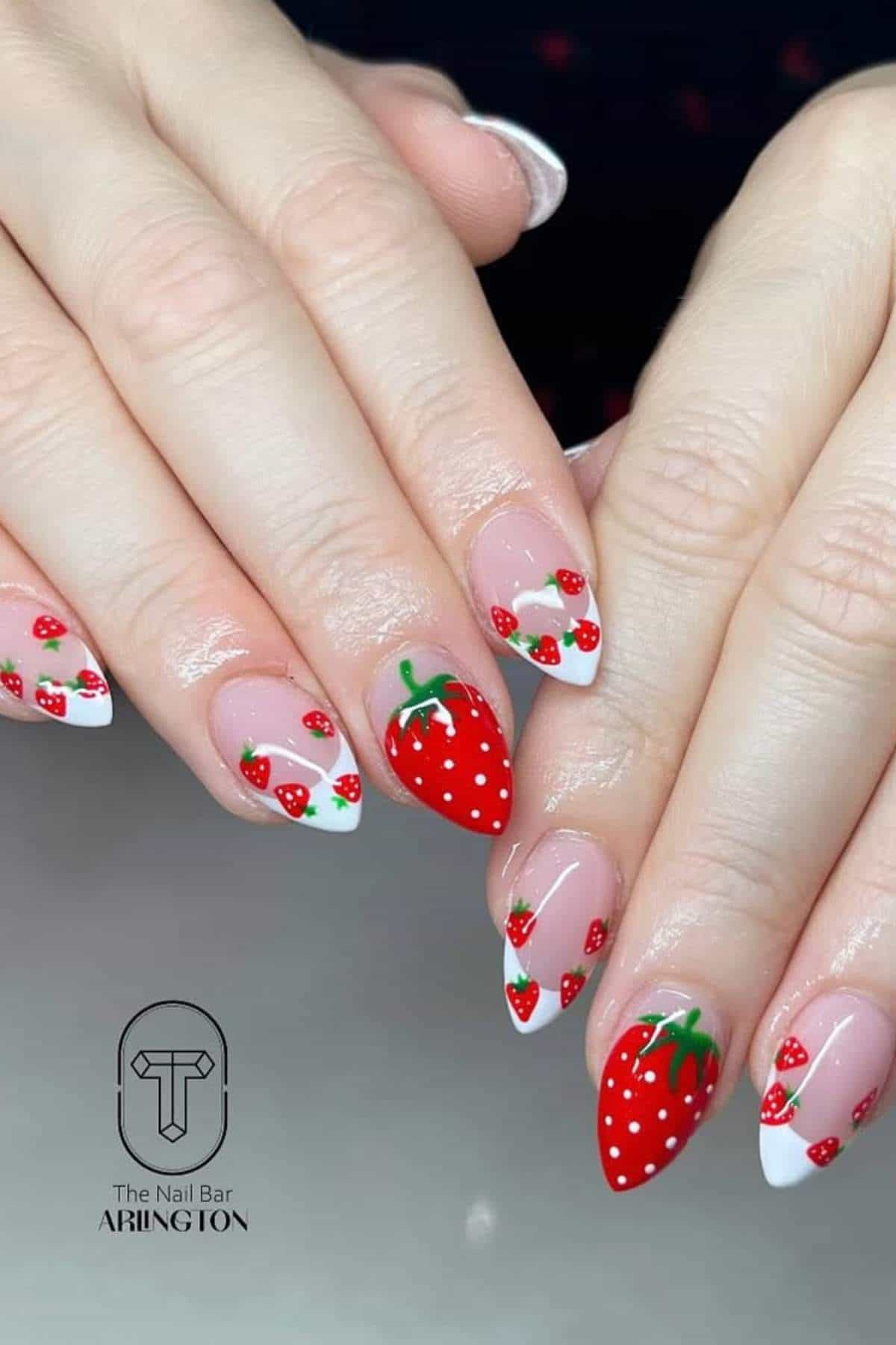 Hands with almond-shaped nails featuring strawberry-themed nail art, including red strawberries with green stems and white tips, photographed on a plain background. Logo: The Nail Bar Arlington.