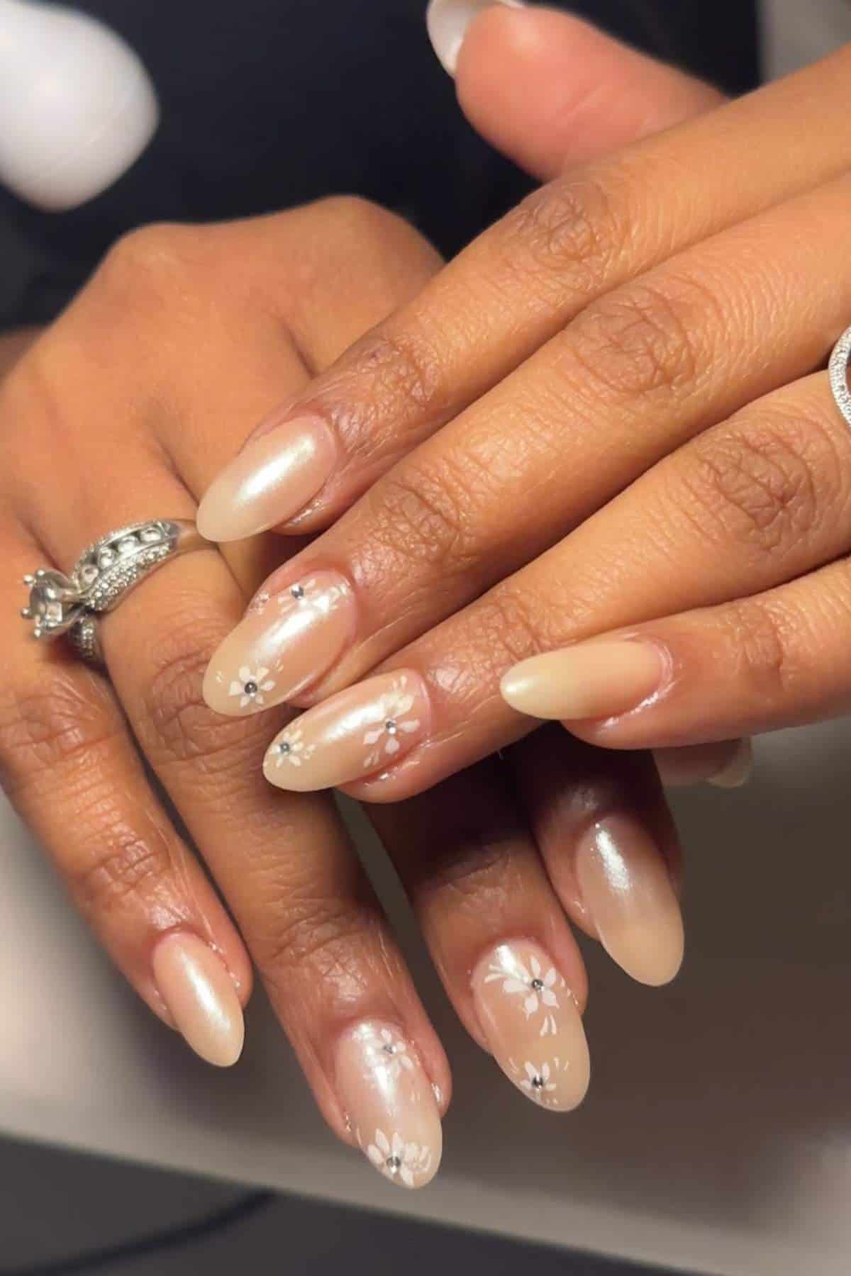 Hands with long, almond-shaped nude nails decorated with small white floral designs. The person is wearing silver rings on their fingers.