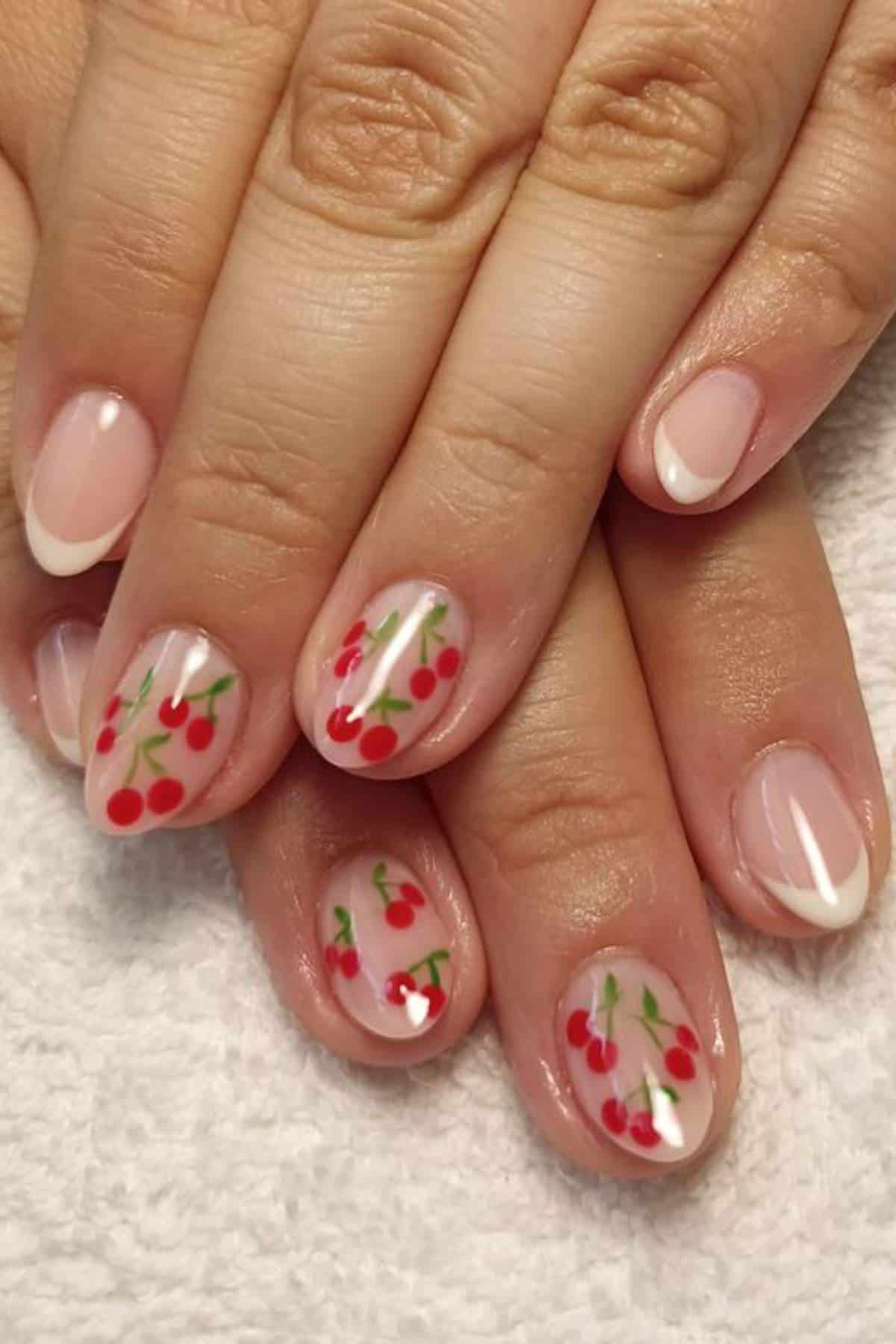 Hands with short, rounded nails. Some nails have French tips and others feature red cherry designs with green stems on a nude base. Nails rest on a white textured surface.