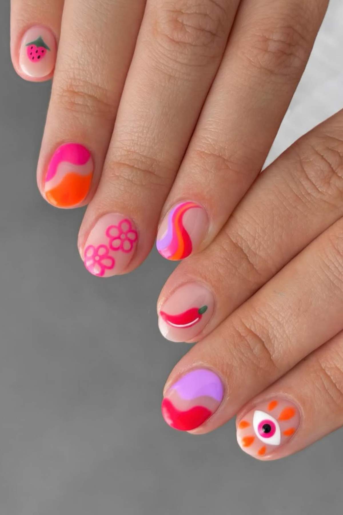 A hand with colorful nail art featuring abstract patterns, a watermelon slice, flowers, a chili pepper, and an eye design on different nails.