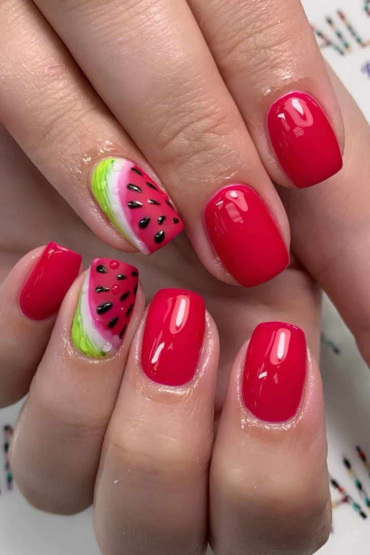 Close-up of hands with glossy red nails; one nail on each hand features a detailed watermelon design with green, pink, and black accents.