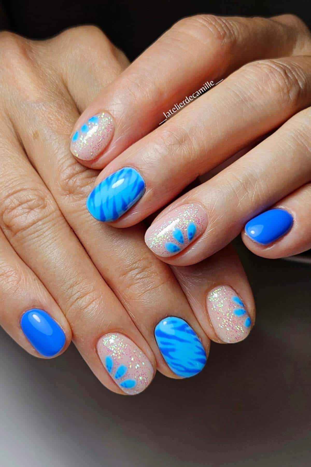 Hands with nails painted in bright blue, shimmer, and clear polish with blue petal and wave designs.