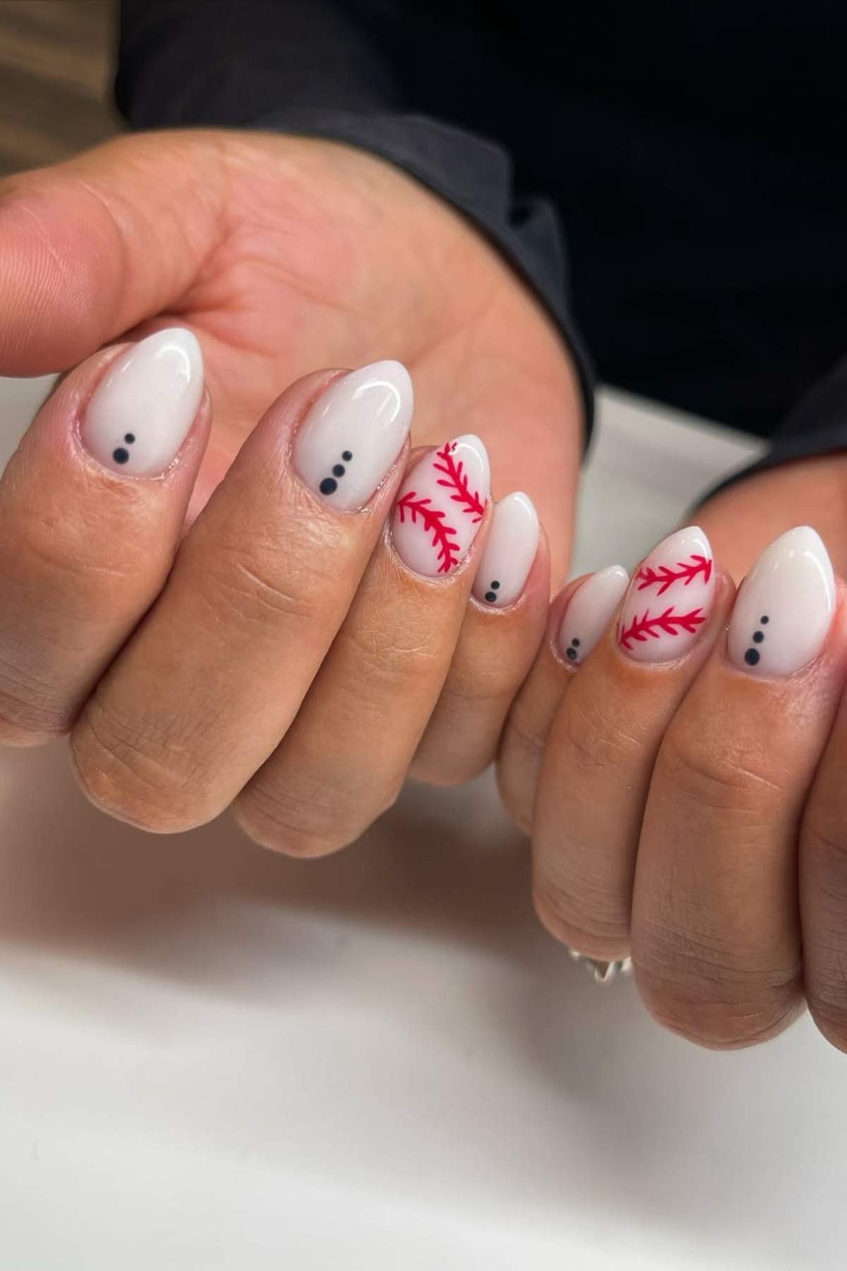 Hands with almond-shaped nails featuring a baseball stitch design on some nails and three black dots on others, all over a pale nude base.