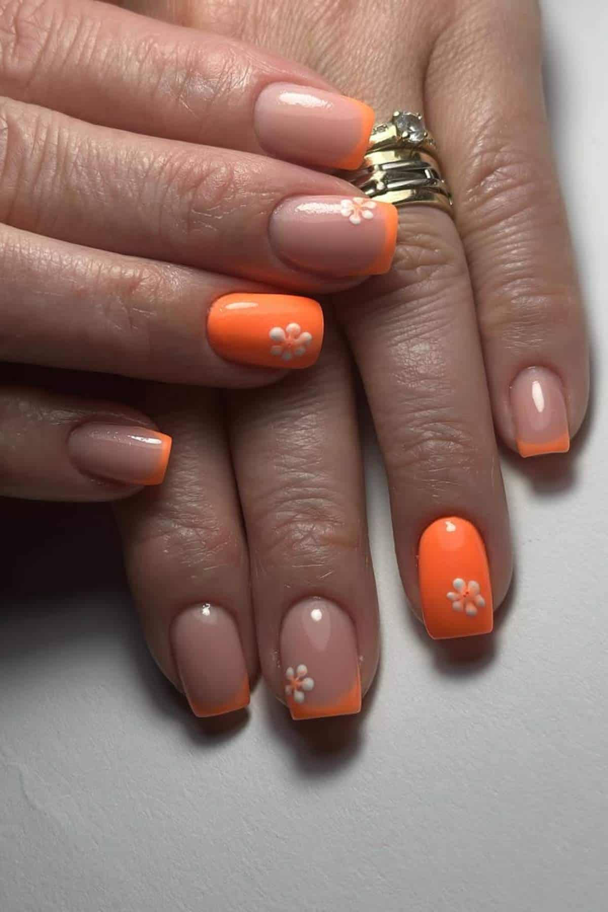 A hand with short square nails featuring a mix of nude and bright orange polish, some with small white flower designs, and wearing gold rings.