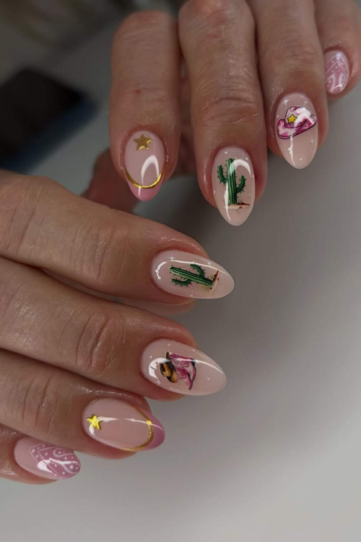 A close-up of hands with almond-shaped nails featuring desert-themed nail art, including cacti, cowgirl hats, boots, stars, and pink bandana patterns on a nude background.