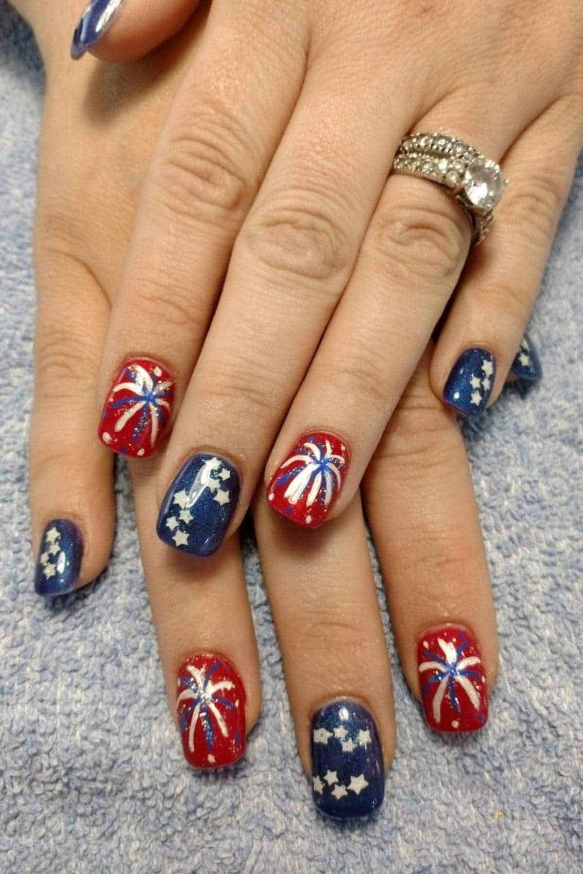 A person's hands display red and blue nails with white star and firework designs, and a diamond ring on one finger.