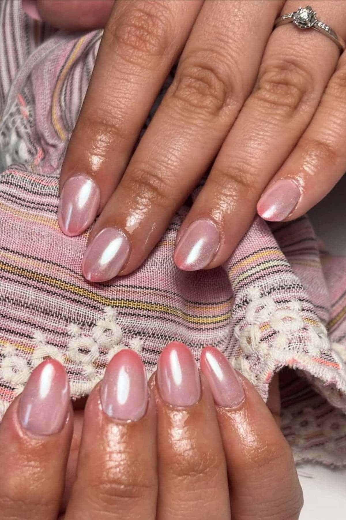 Close-up of hands with almond-shaped nails painted in a shiny, pearlescent light pink polish, holding a patterned pink fabric.