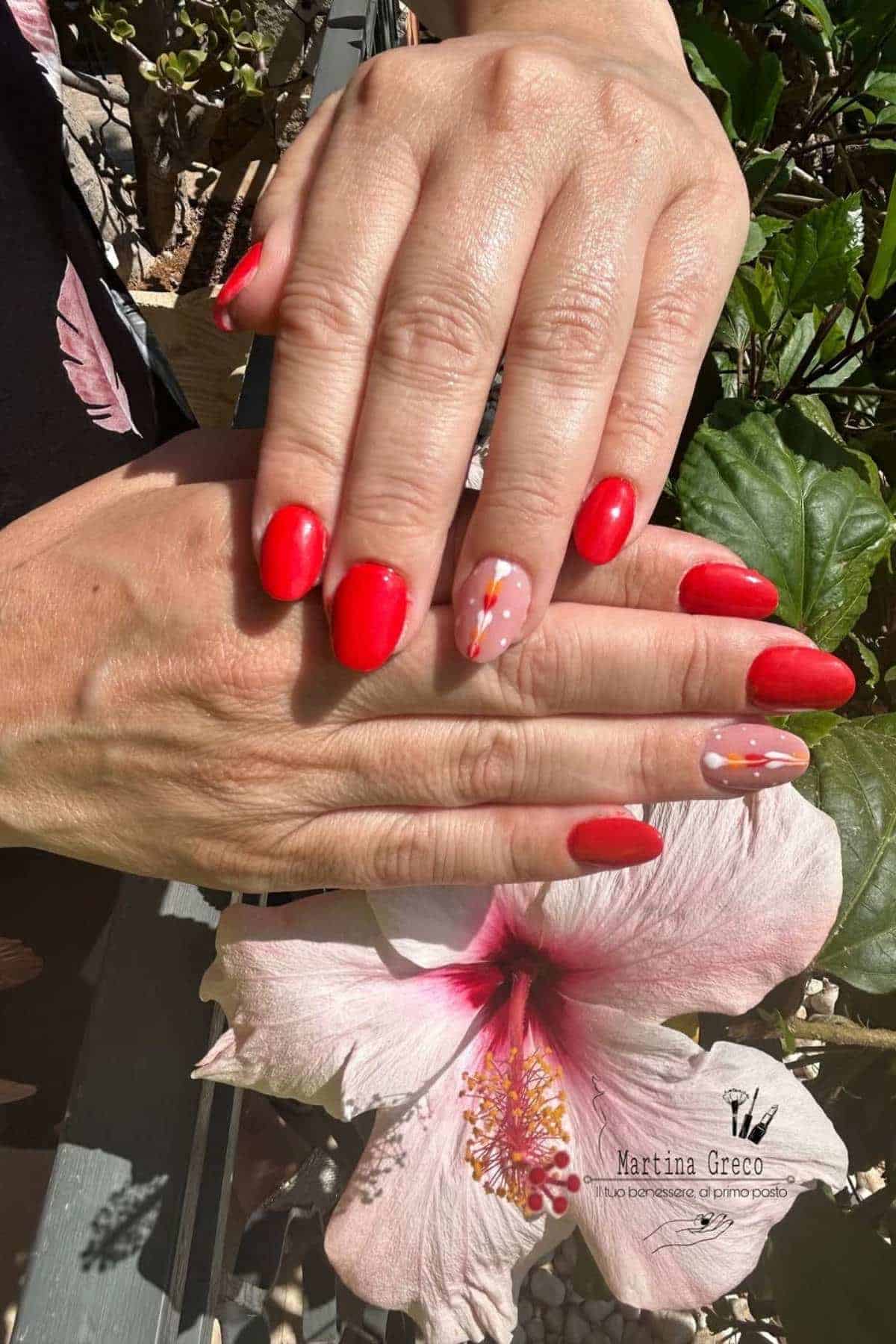 Hands with bright red nail polish and floral nail art accent finger, displayed above a pink hibiscus flower.