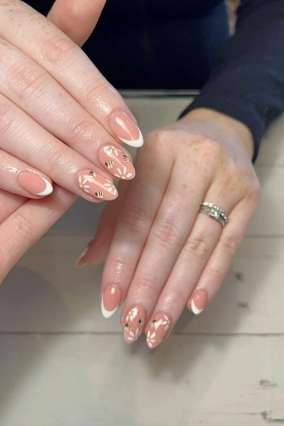 A hand with almond-shaped nails painted in a nude color with white tips and gold leaf nail art accents, resting on a light wooden surface.