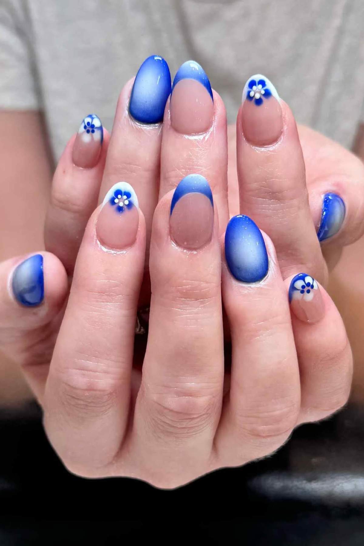 Hands with almond-shaped nails featuring blue and white ombre tips, French tips, and small blue and white flower designs on some nails.