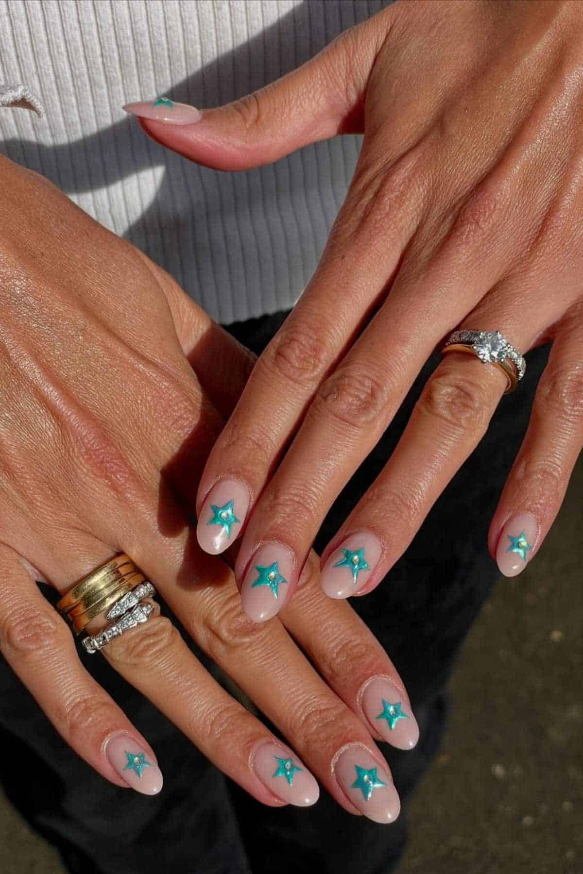 Hands with manicured nails featuring teal star designs on a nude base, wearing gold and silver rings, against a neutral background.