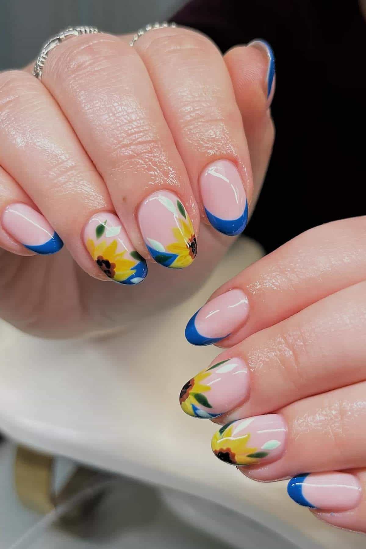 Hands with manicured nails featuring blue French tips and sunflower designs on a light pink base.
