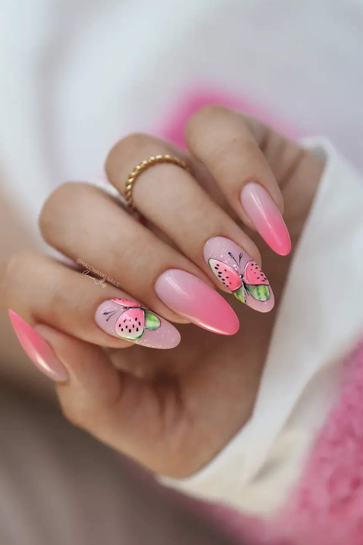 A hand with long, almond-shaped pink ombre nails, featuring a butterfly design on the ring and middle fingernails.