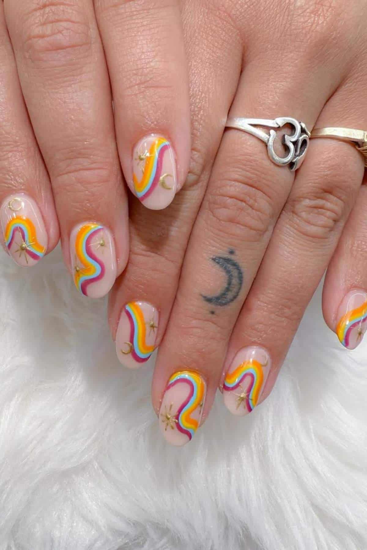 A hand with rainbow swirl nail art, gold star and ring accents, and a crescent moon tattoo on the finger, resting on a white textured surface.