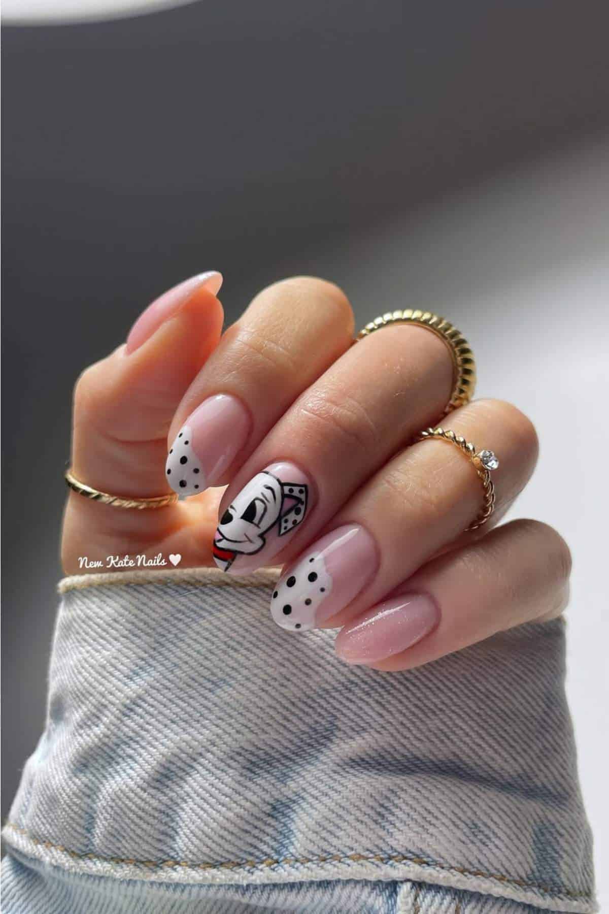 A hand with manicured nails featuring pink polish, black dots, and one accent nail with abstract face art. The person wears gold rings and a light denim jacket.