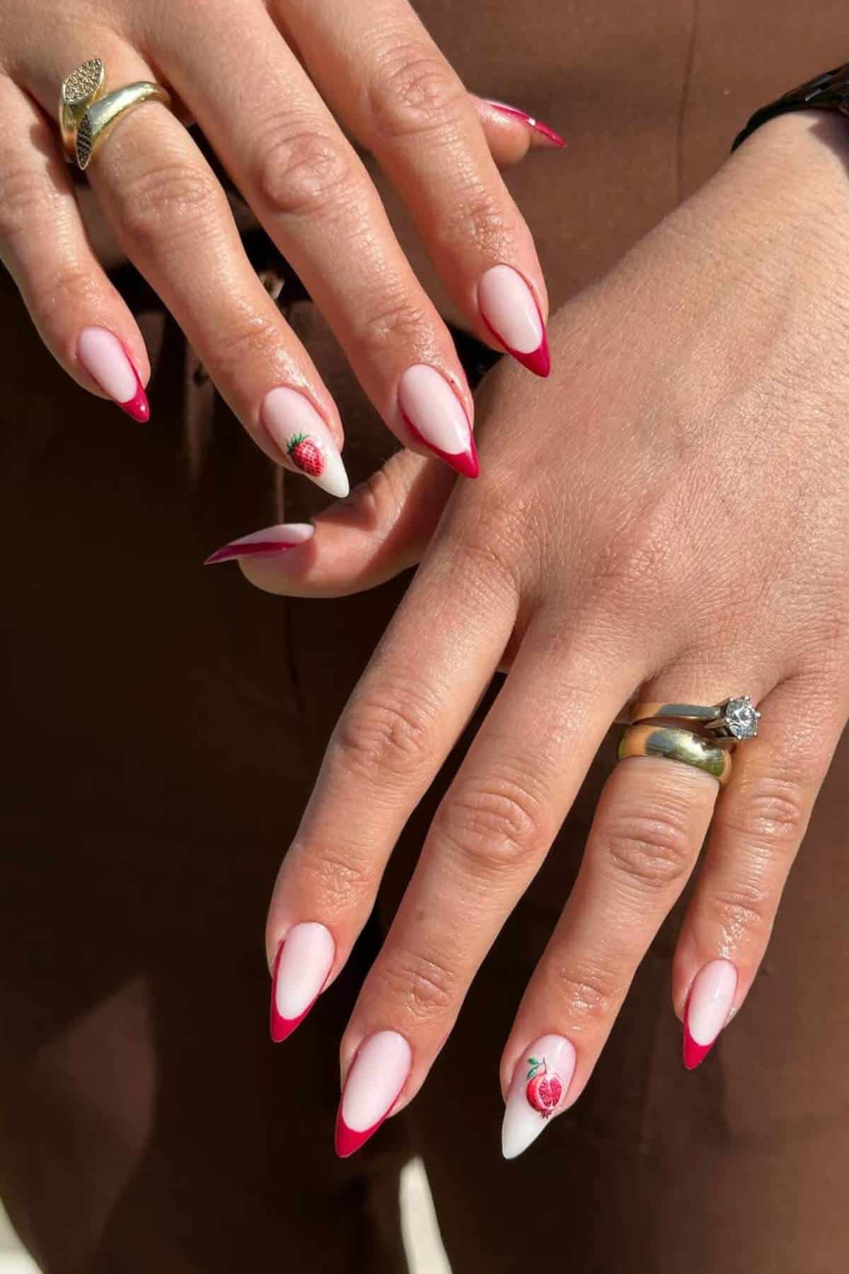 A close-up of hands with almond-shaped nails featuring pink bases, red French tips, and small rose designs on two accent nails. The person is wearing gold rings.