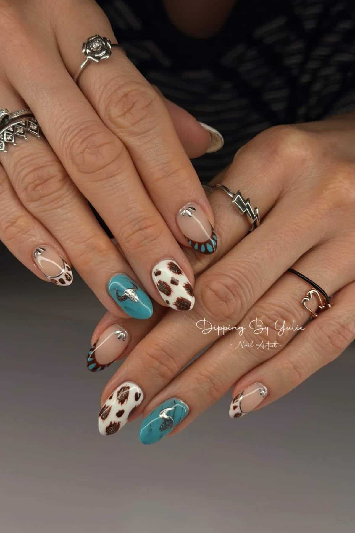 Hands with long almond-shaped nails featuring a mix of cow print, turquoise, and nude polish designs, accented with silver nail charms and geometric rings.