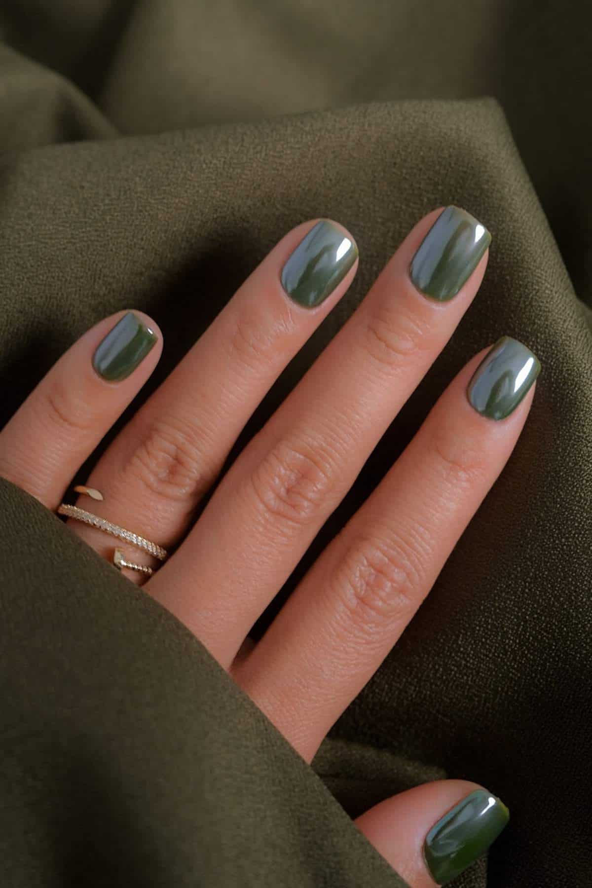 A hand with short, glossy olive green nails resting on an olive green fabric, wearing thin gold rings on the ring finger.