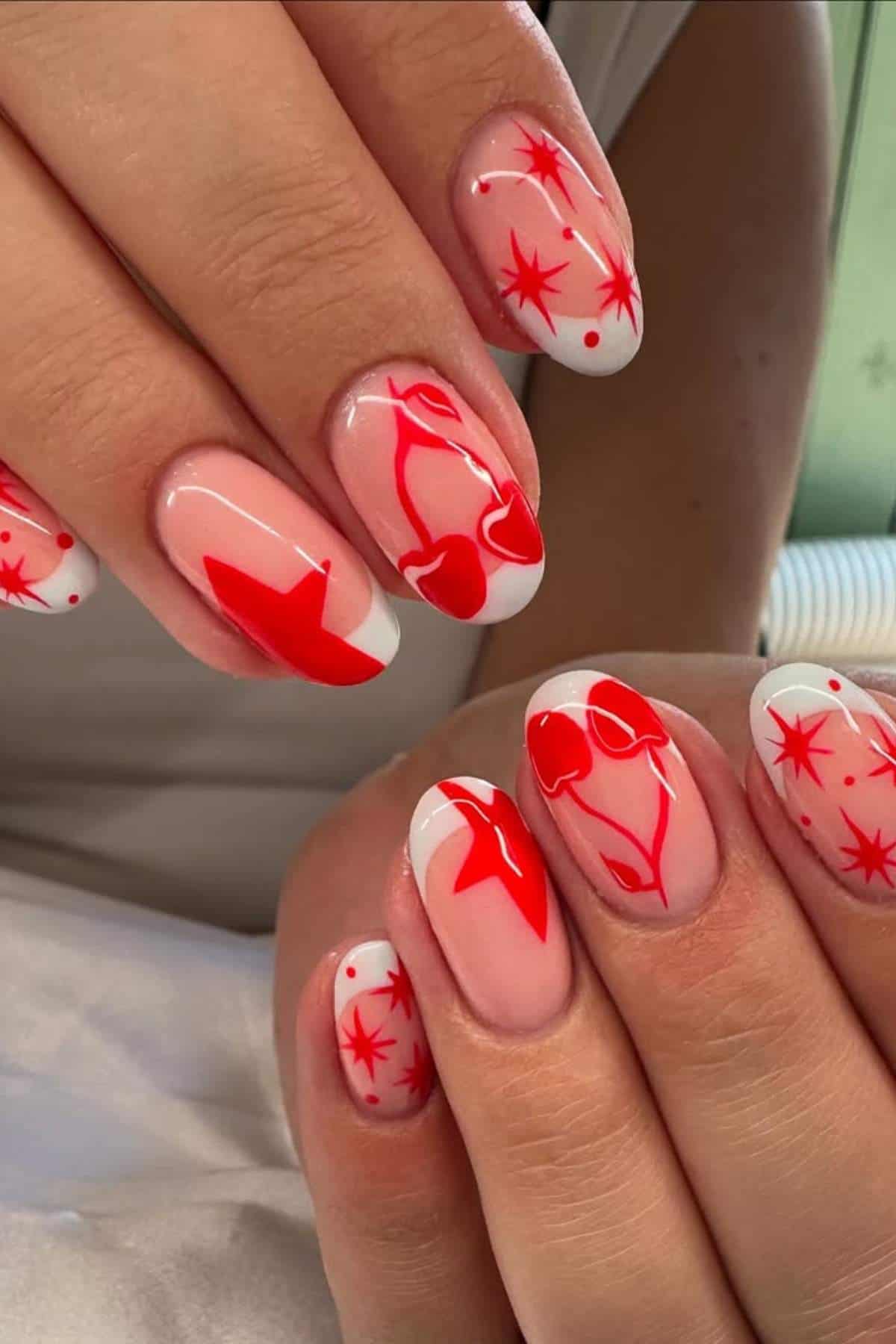 Close-up of hands with red abstract nail art on a light pink base, featuring stars, shapes, and outline designs on short, rounded nails.
