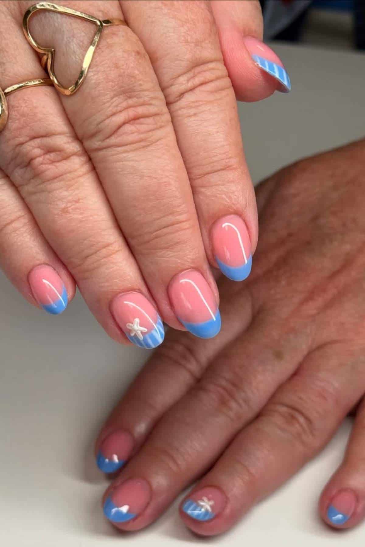 Hands with manicured nails featuring light blue French tips, white accents, and small starfish designs on a nude pink base. One hand has a gold heart-shaped ring.