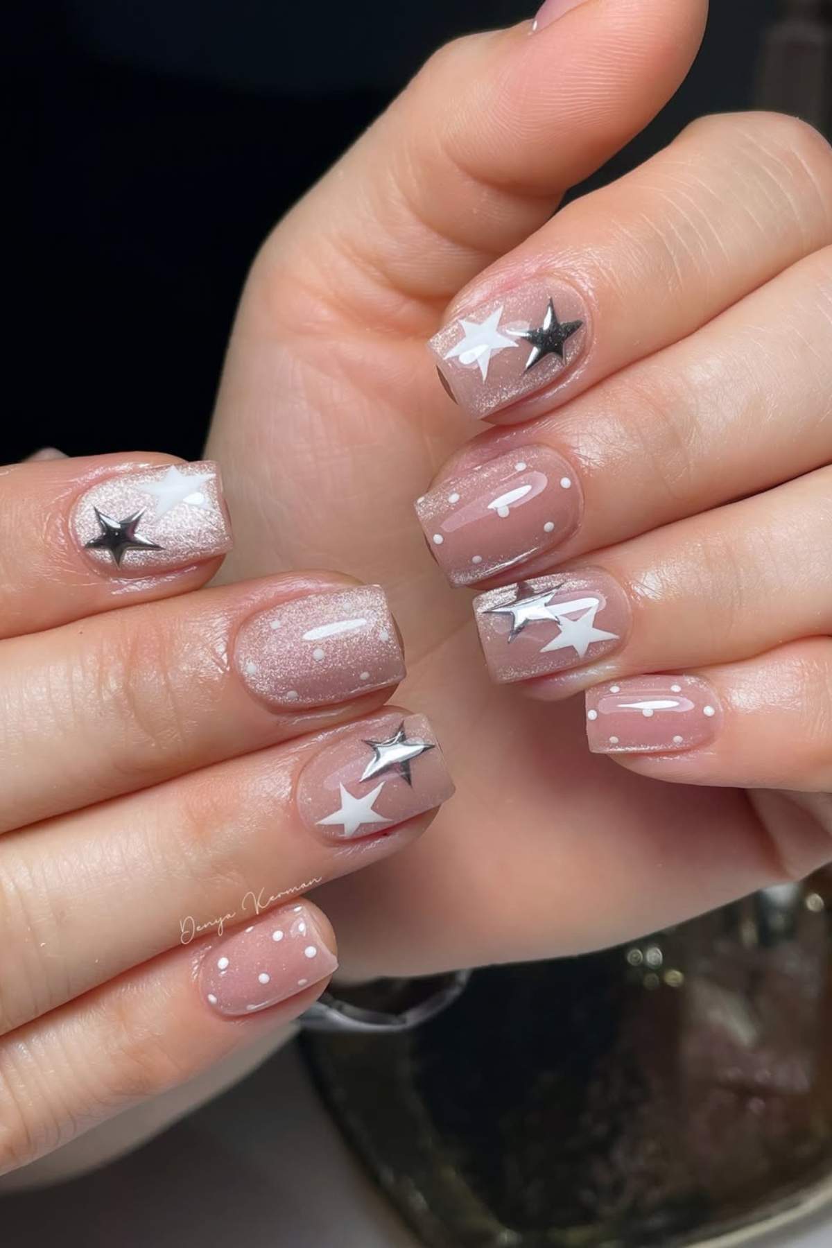 Hands with short, square-shaped nails featuring nude polish, white and black star designs, white dots, and glossy finish.