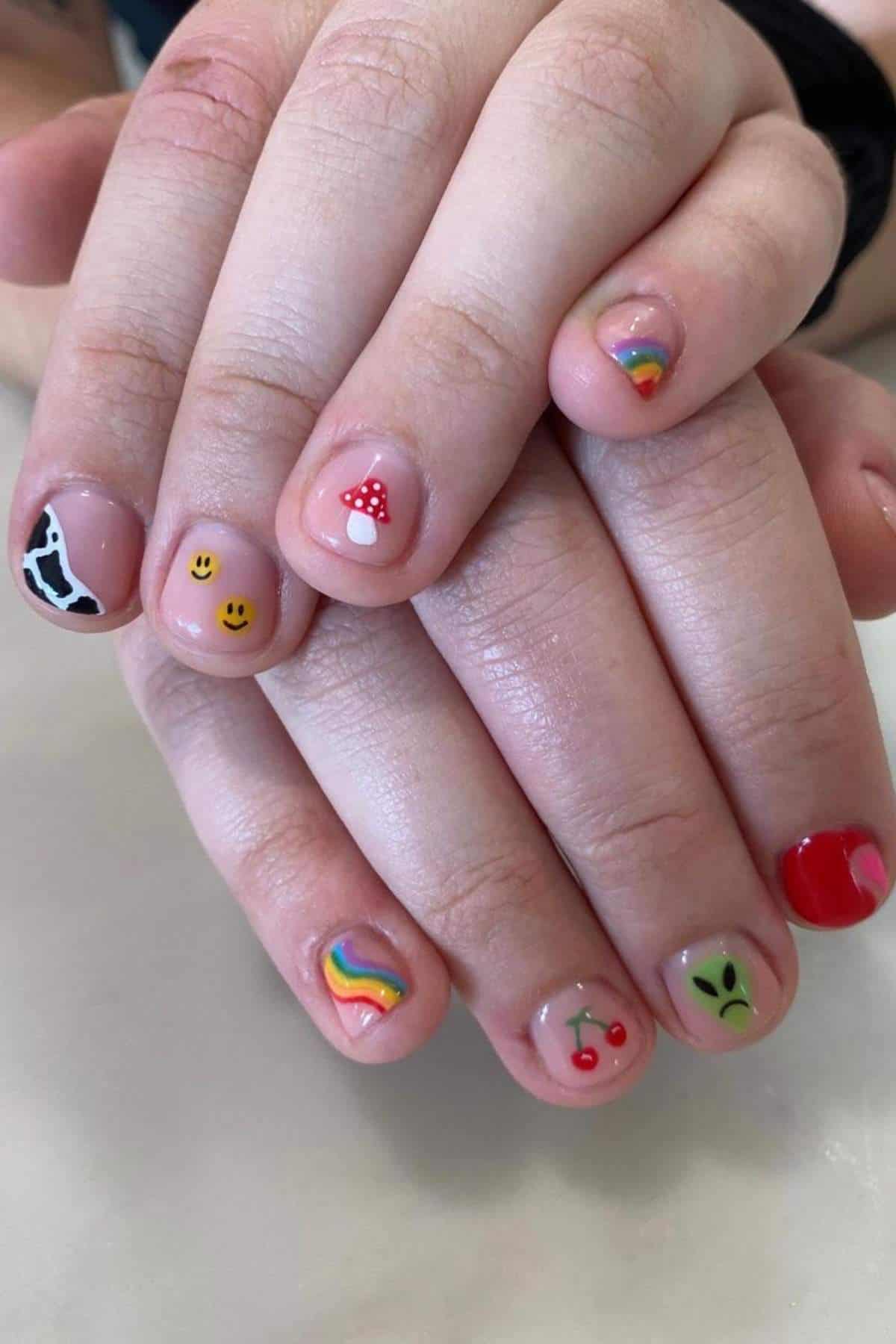 Two hands with short nails, each nail painted with a different design including a cow print, smiley faces, mushroom, rainbows, cherries, alien face, and a red heart.