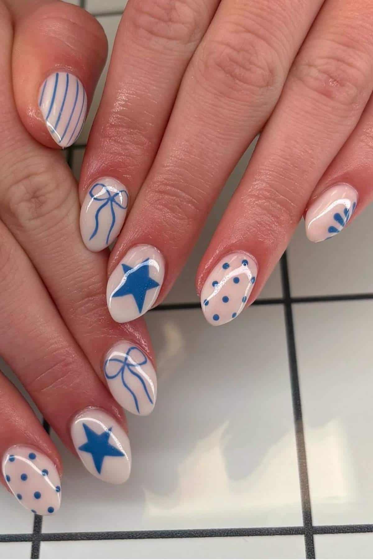 Hands with almond-shaped nails painted in a nude base, decorated with blue lines, stars, bows, and polka dot designs, displayed over a white tiled surface.