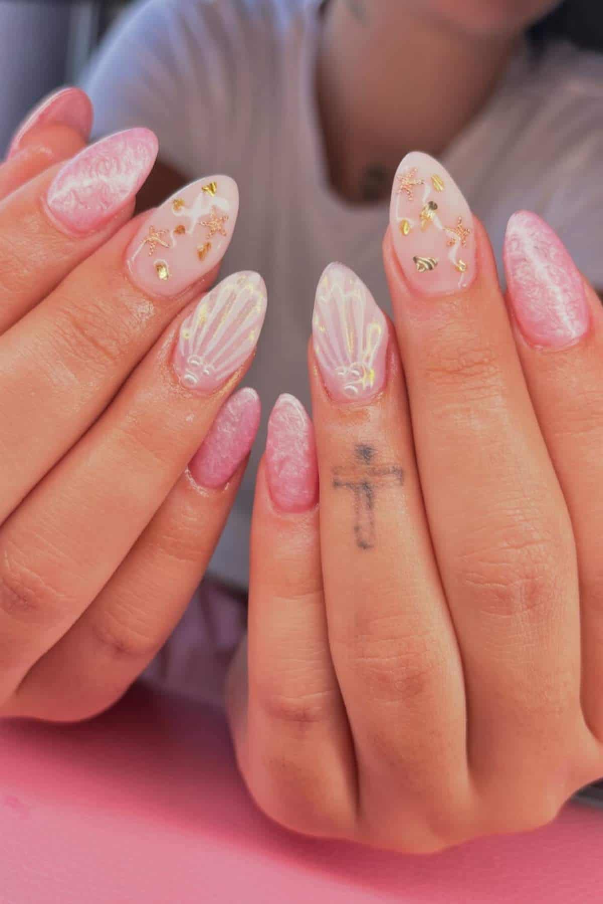 A person with long, almond-shaped pink and white manicured nails featuring gold accents and shell designs, with a small cross tattoo on their middle finger.