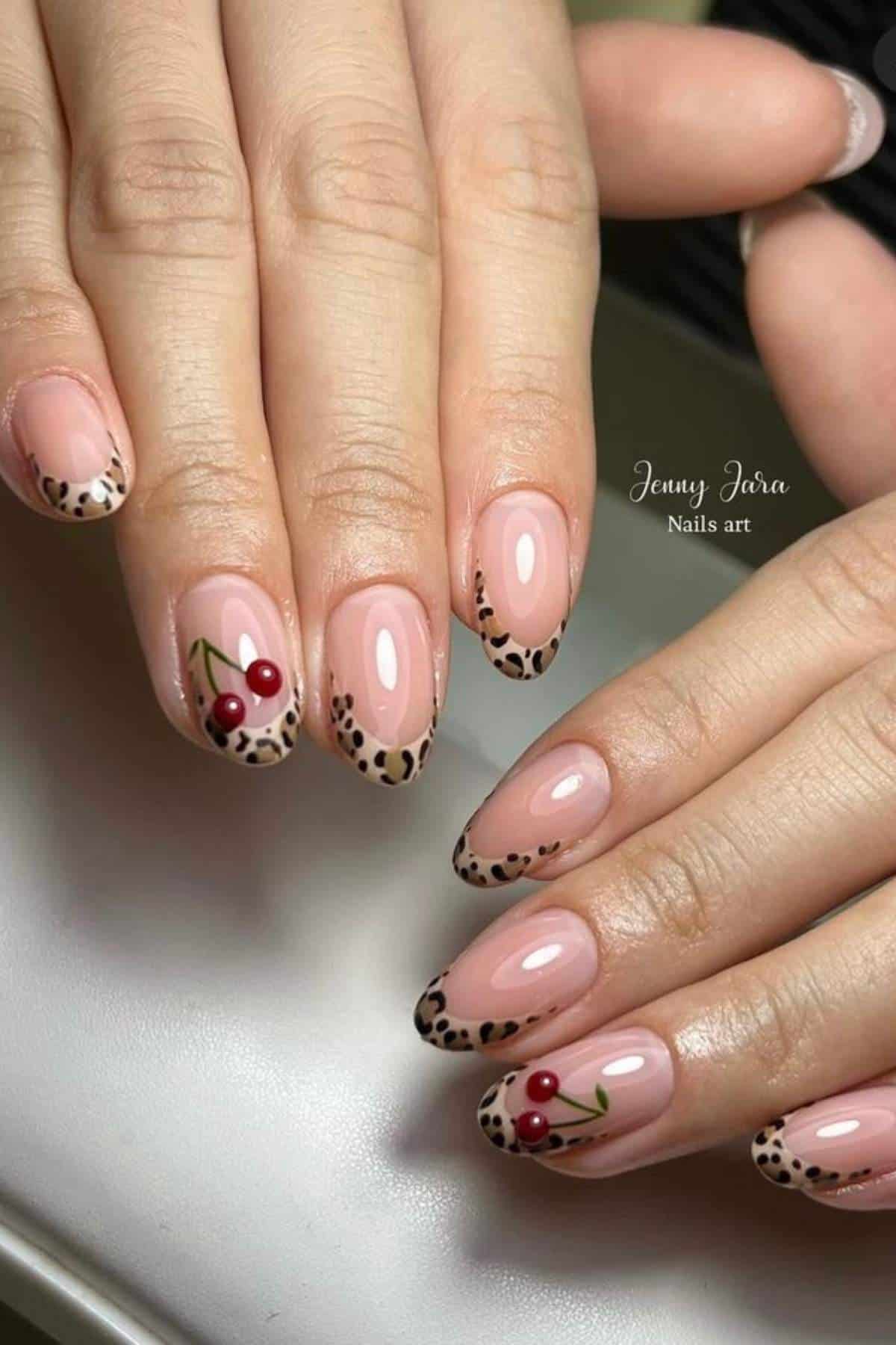 Hands with almond-shaped nails featuring nude polish, cheetah print tips, and two accent nails with red cherry designs.