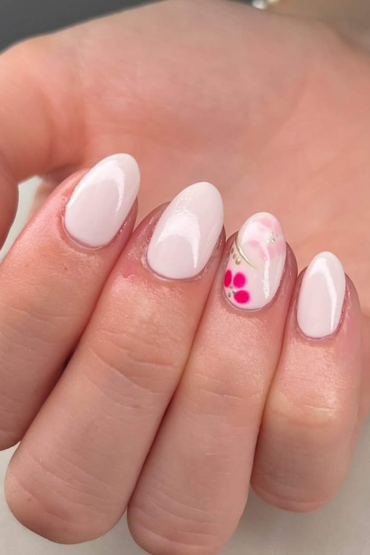 A hand with short, oval-shaped nails painted pale pink; the ring fingernail features a floral design in pink and white.