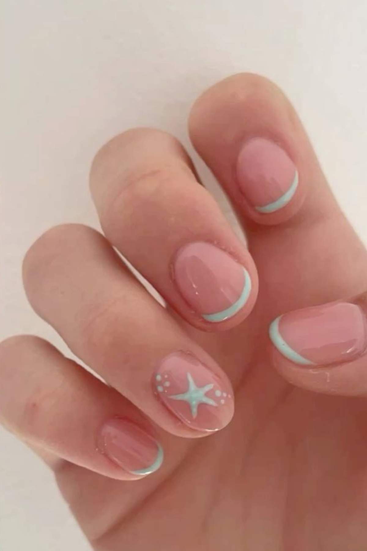 A hand with short, oval-shaped nails painted light pink, featuring light blue French tips and a blue starfish design with white dots on the ring finger.