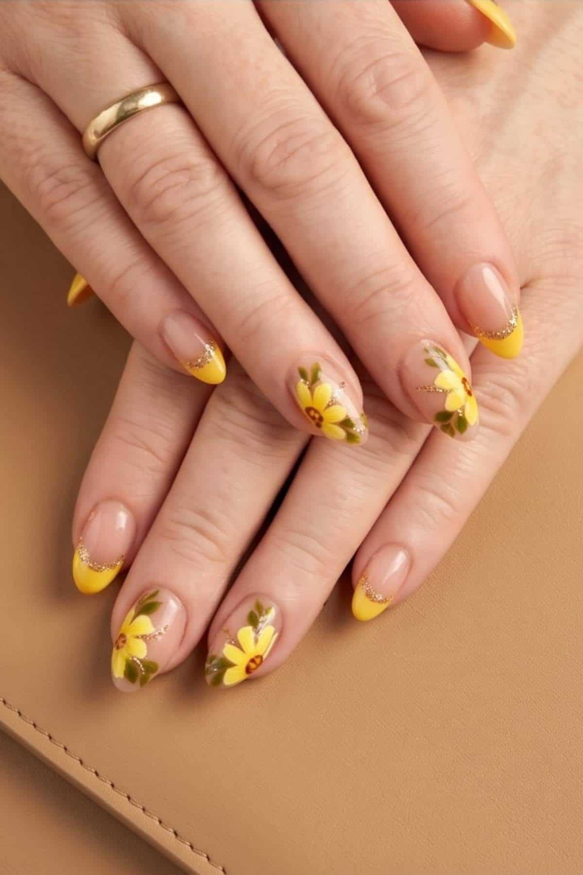 Hands with yellow French tip manicure featuring yellow flower designs and gold accents, resting on a tan surface. A gold ring is worn on one finger.
