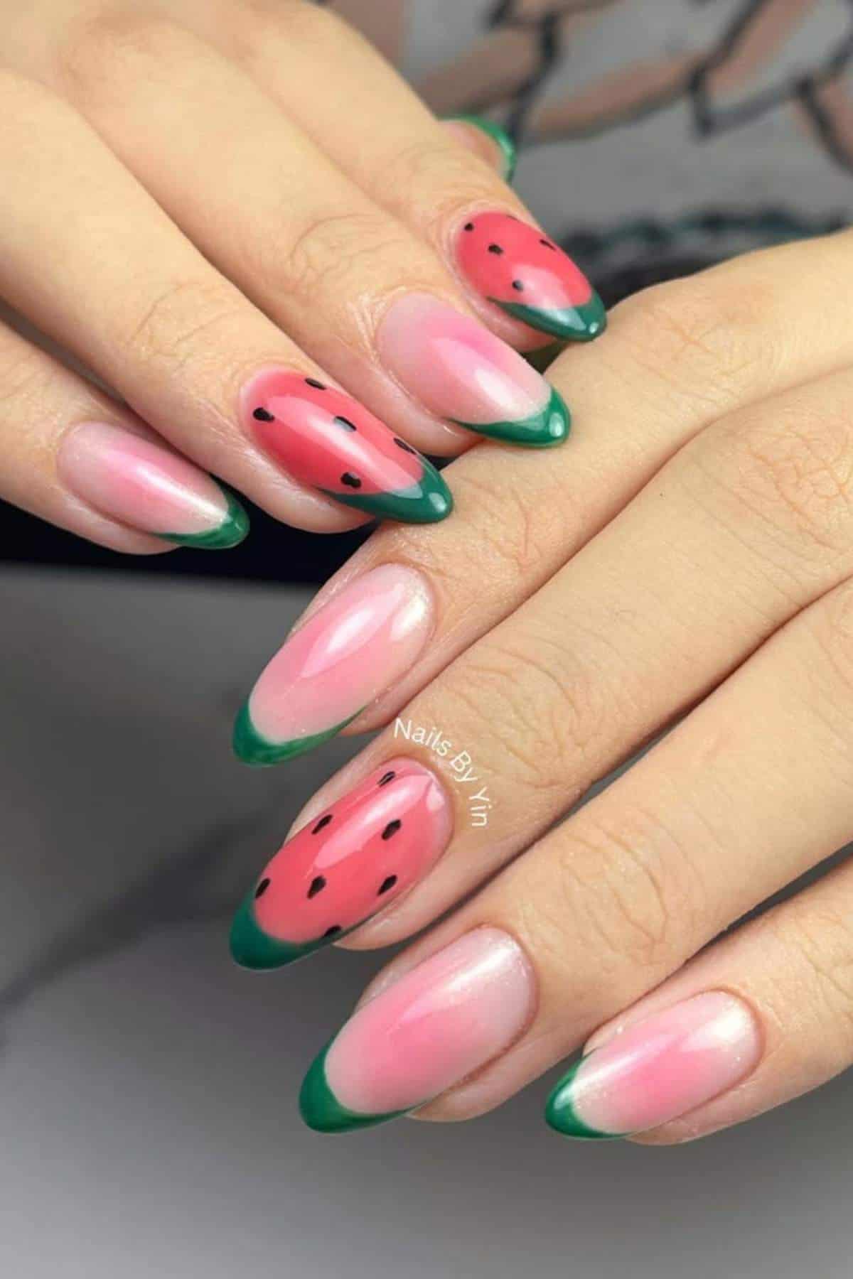 Hands with almond-shaped nails featuring a watermelon-inspired design; some nails are pink with green tips and black dots, while others have simple green French tips.