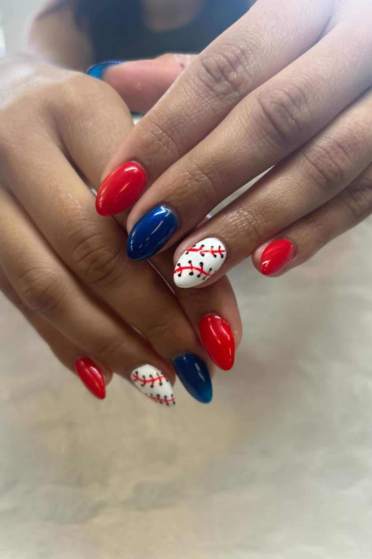 Hands with manicured nails painted in red, blue, and white, with several nails featuring a design resembling baseball stitching.