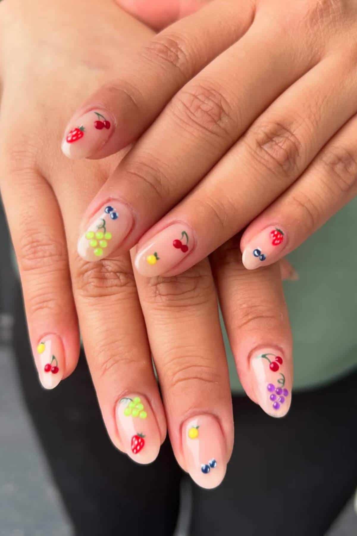 Hands with long, almond-shaped nails featuring colorful fruit designs like cherries, strawberries, and grapes on a natural base.