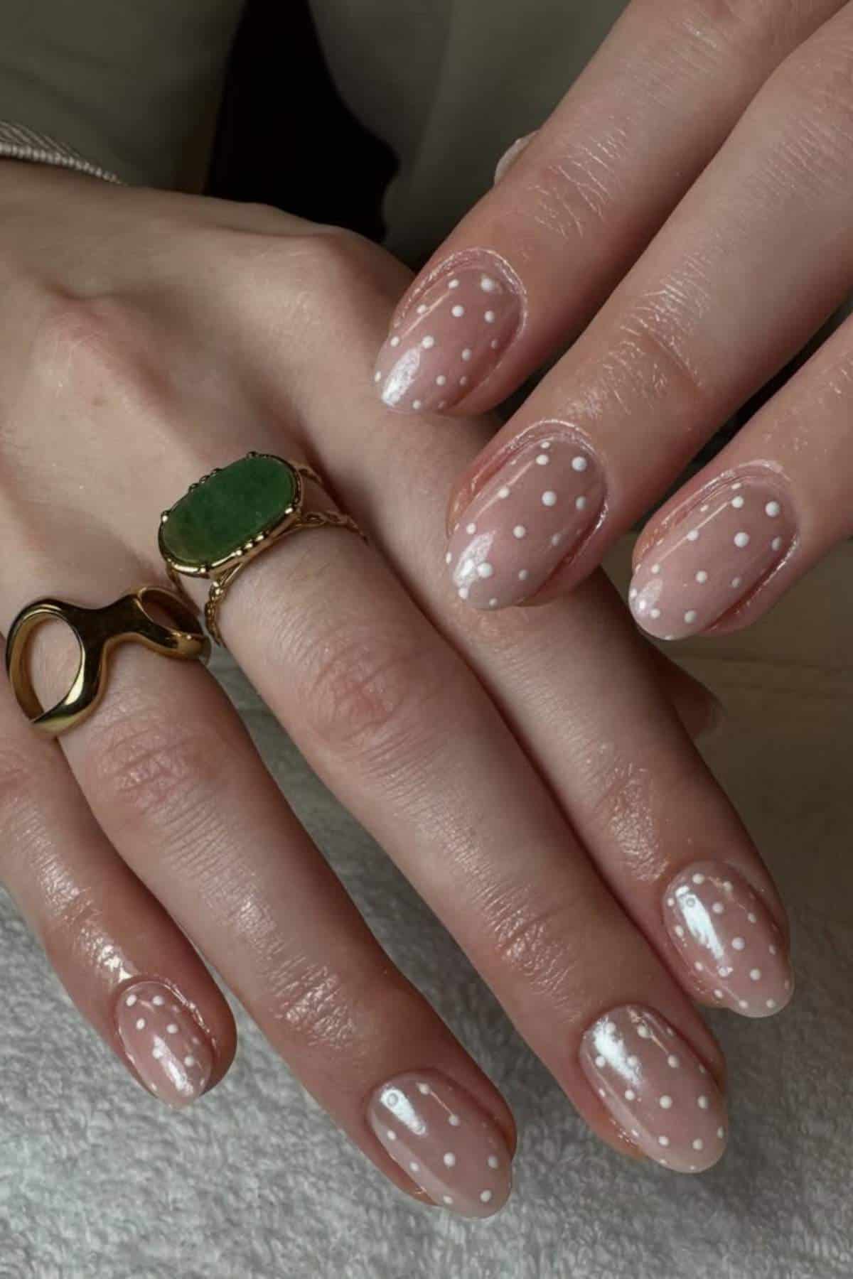 Close-up of hands with nude nails featuring white polka dot nail art, wearing a green stone ring and a gold abstract-shaped ring.