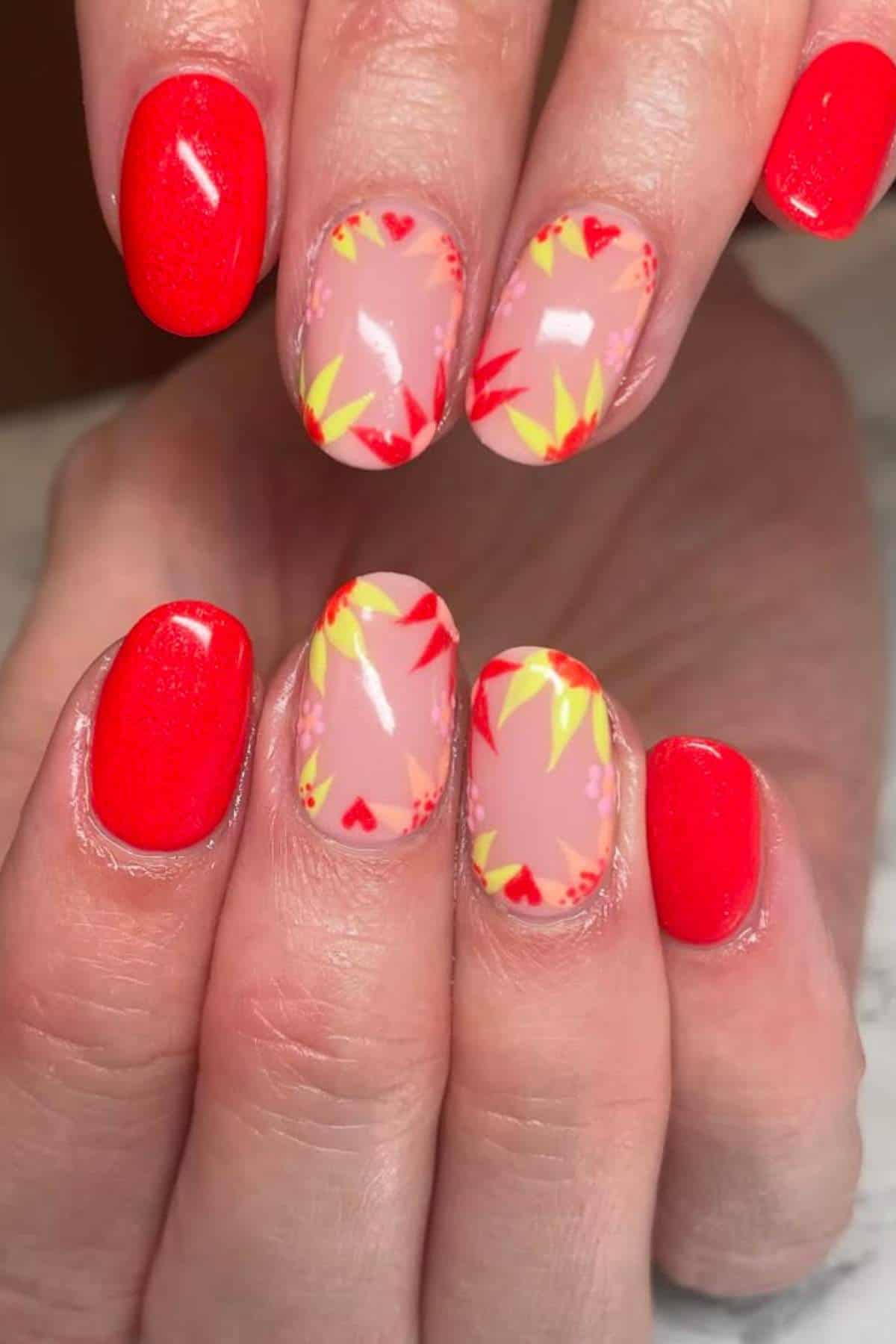 Close-up of hands with red painted nails; two nails on each hand feature a floral and heart pattern on a nude base with red and yellow accents.