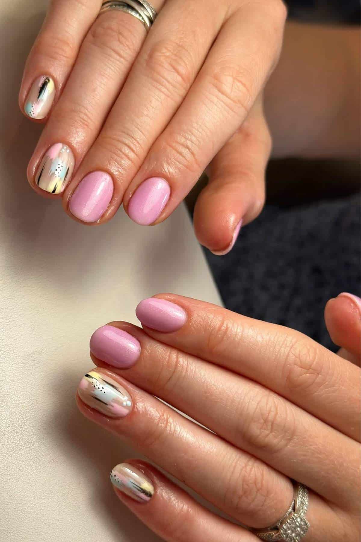 Hands with short, rounded nails featuring pink polish and accent nails with abstract designs in gold, white, and black. The hands display silver rings.