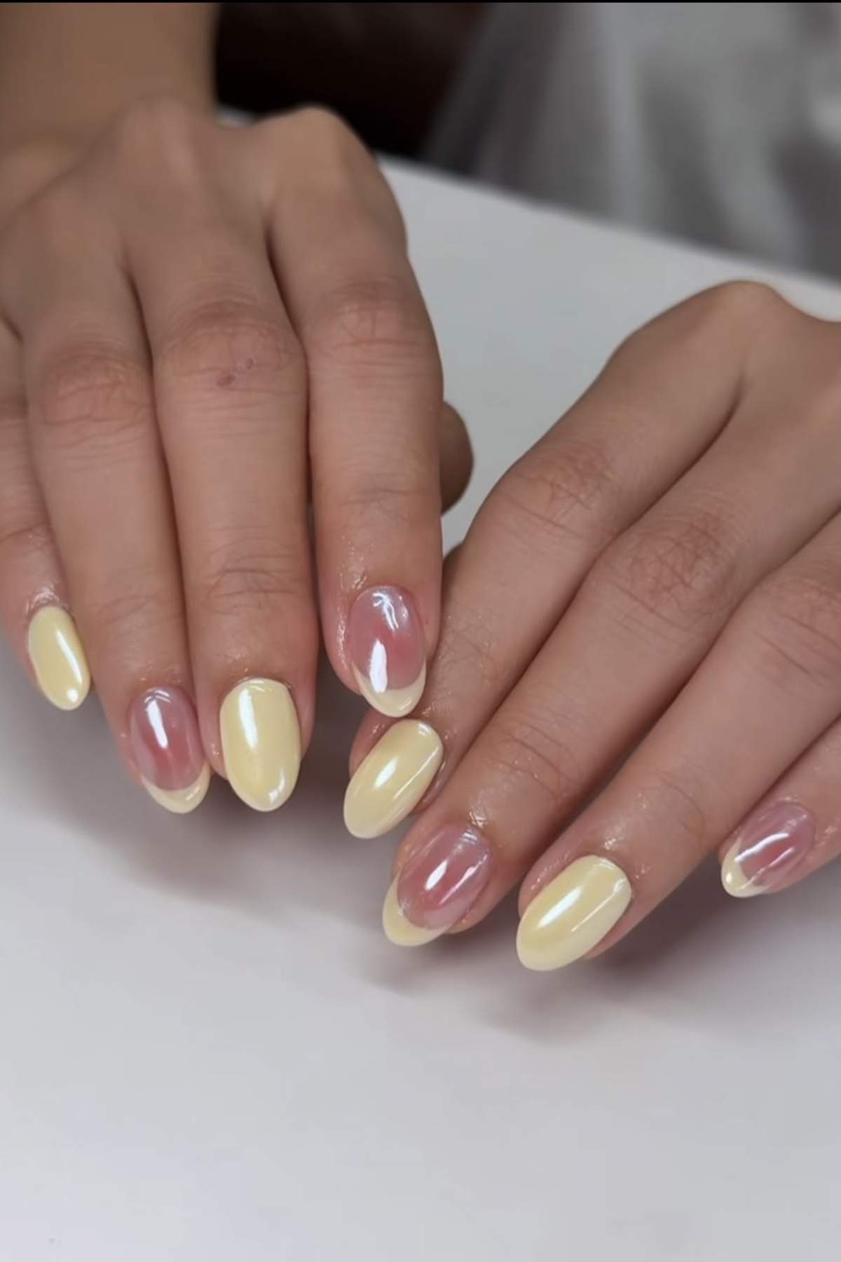Two hands with almond-shaped nails featuring a mix of pastel yellow polish and clear polish with yellow accents on a white surface.