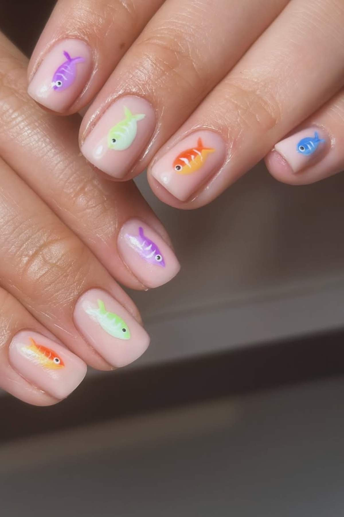 Fingernails painted with a nude base, each nail featuring a small, colorful cartoon fish design in different colors.