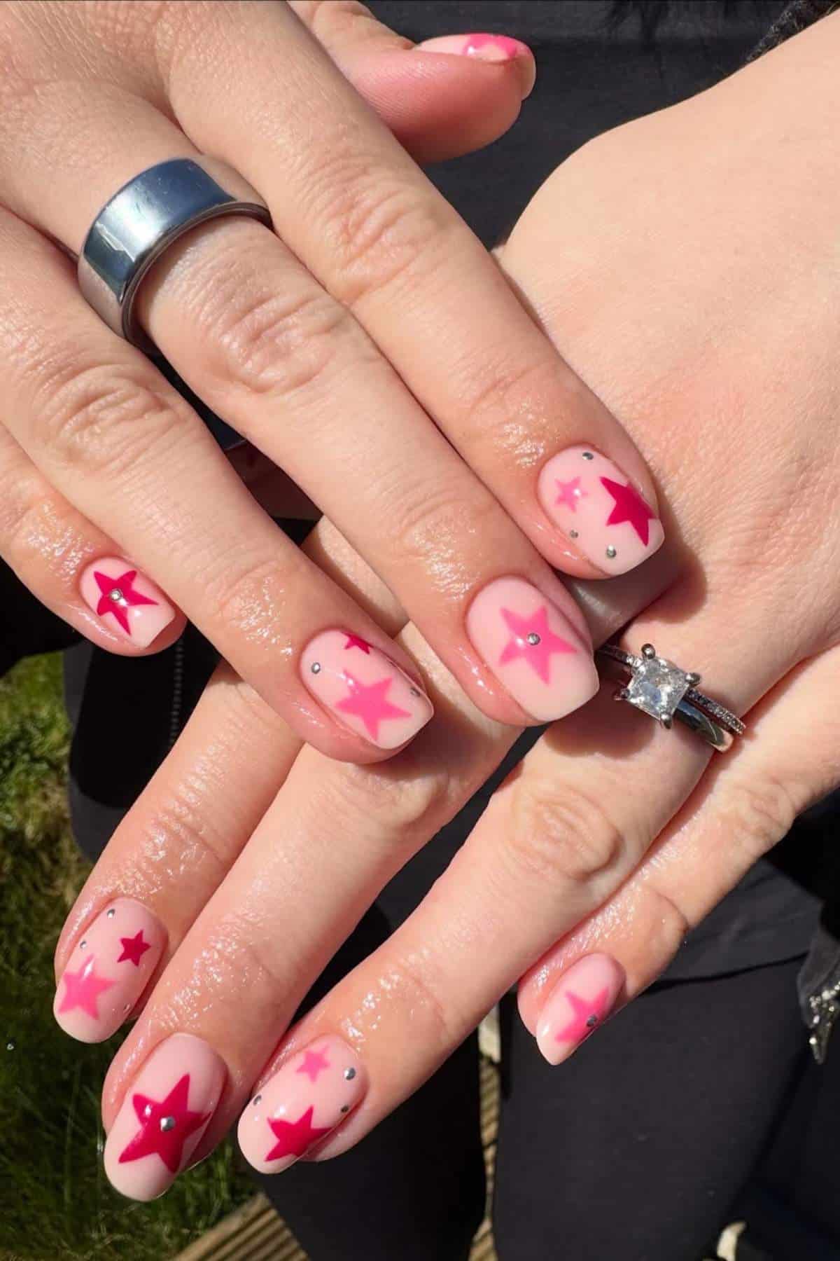 Hands with manicured nails featuring pink and red star designs, small rhinestones, and a silver and diamond ring on the ring finger.