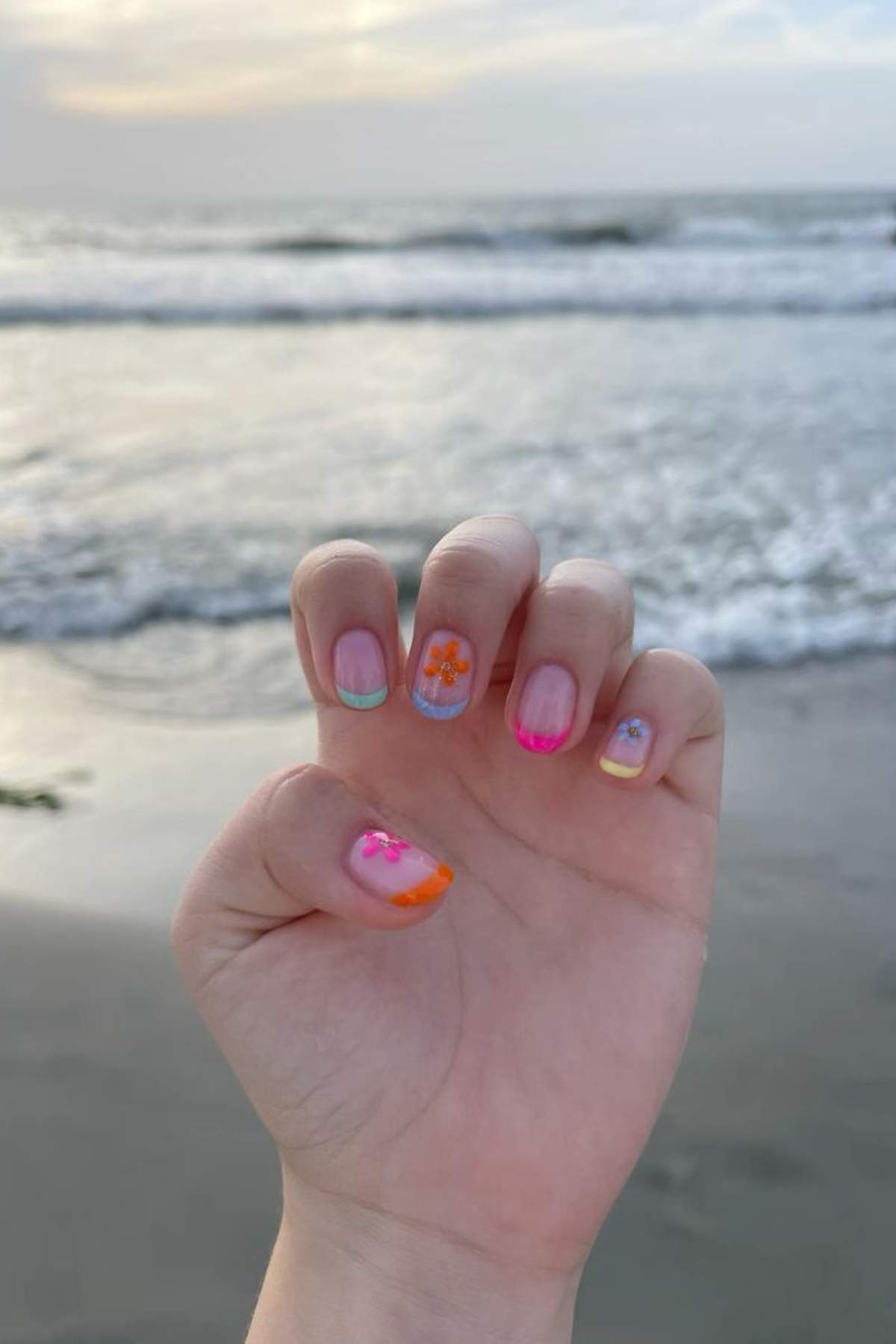 A hand with colorful floral nail art is held up in front of the ocean, with gentle waves and a cloudy sky in the background.