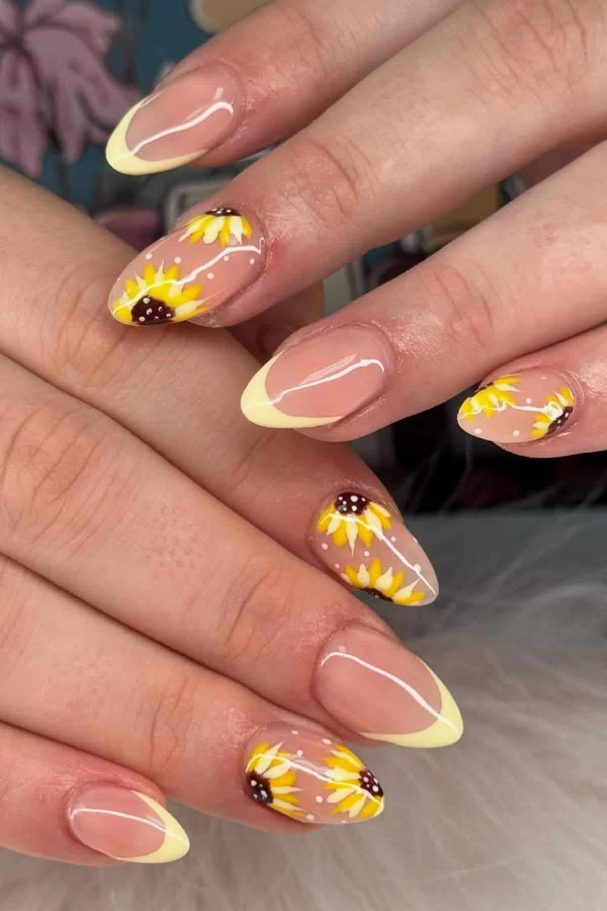 Hands with almond-shaped nails featuring pale yellow French tips and sunflower designs on a nude base.