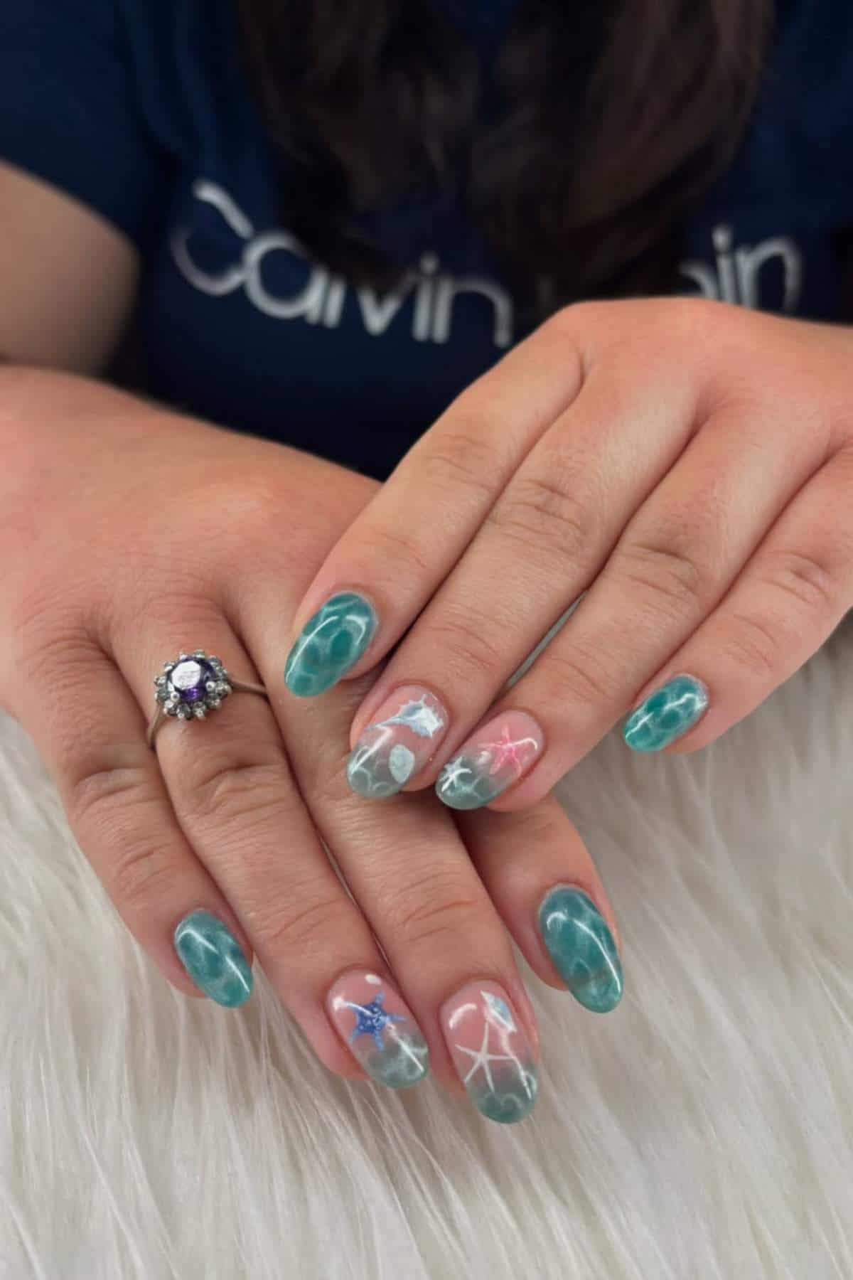 Hands with teal and pink marble nail art featuring white and blue starfish designs, resting on a white textured surface. A ring with a purple stone is worn on one finger.