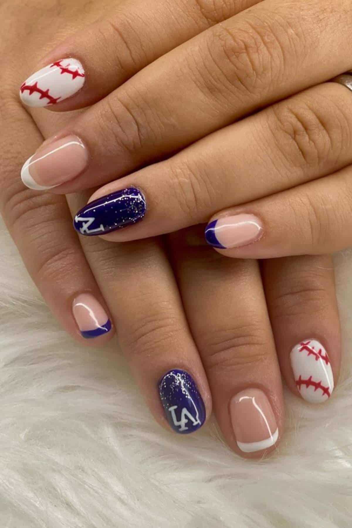 Close-up of hands with manicured nails featuring baseball stitching, French tips, and blue nails with "LA" letters and glitter accents.