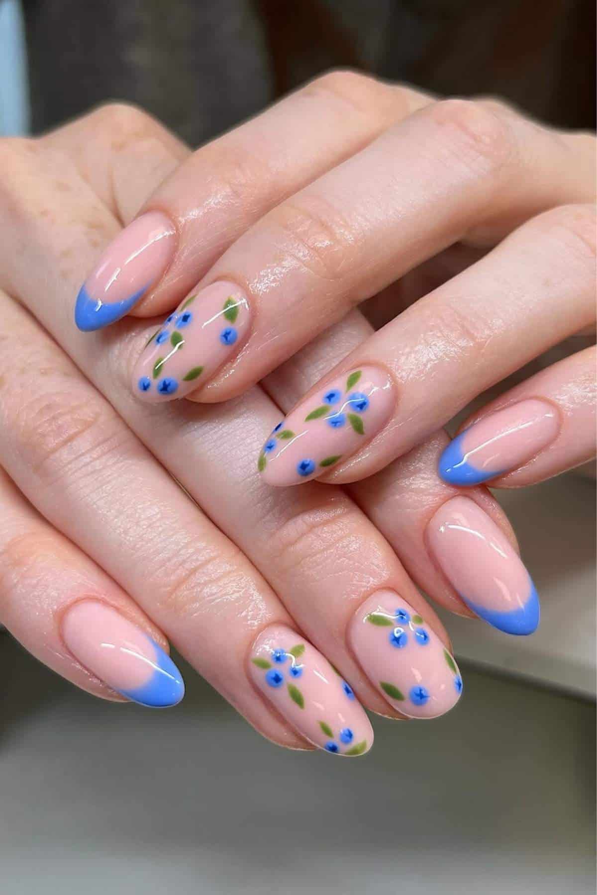 Hands with almond-shaped nails featuring a nude base, blue French tips, and blue flower designs with green leaves on some nails.