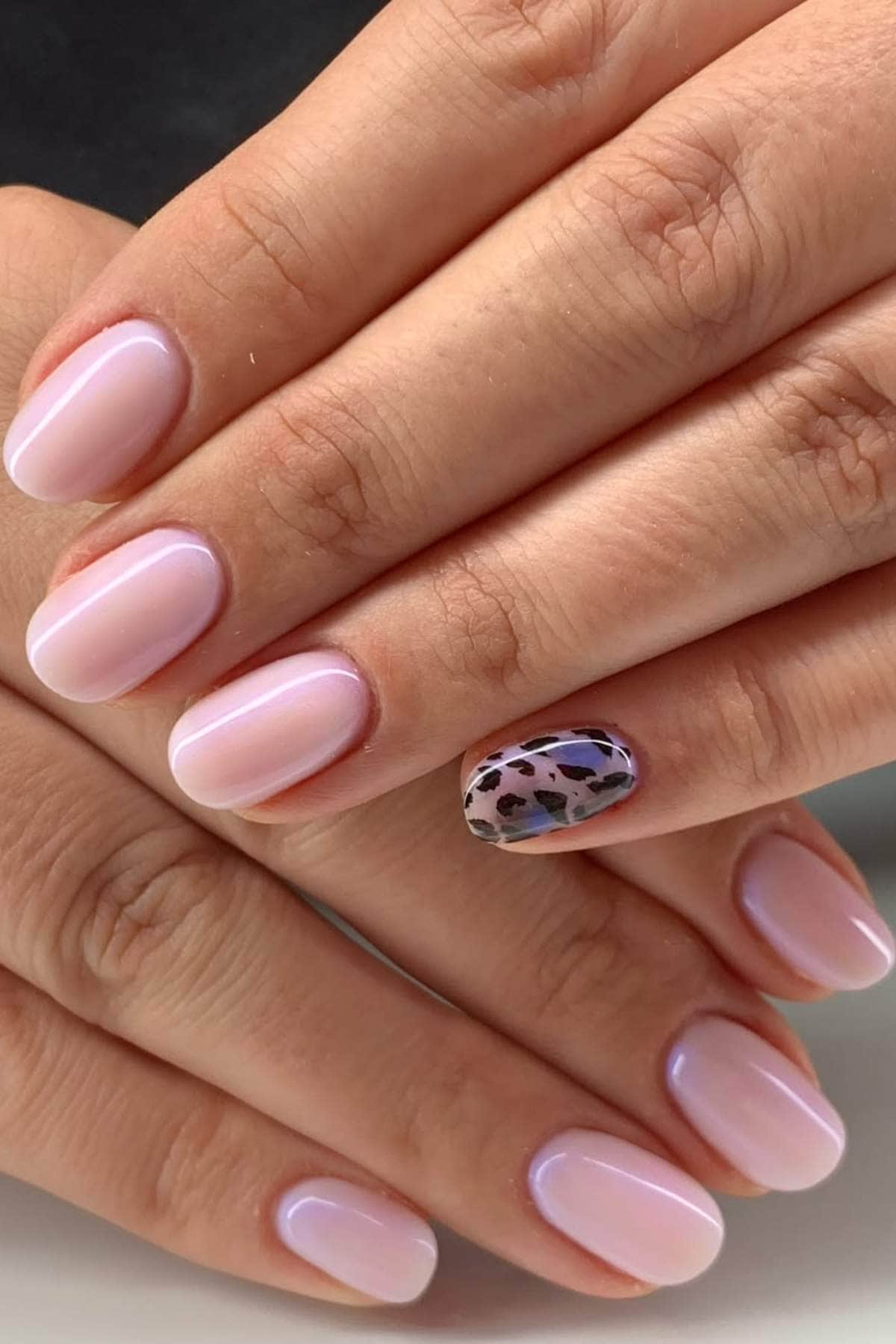 Hands with short, oval-shaped nails painted in a light pink polish; one accent nail features a black and brown leopard print design.