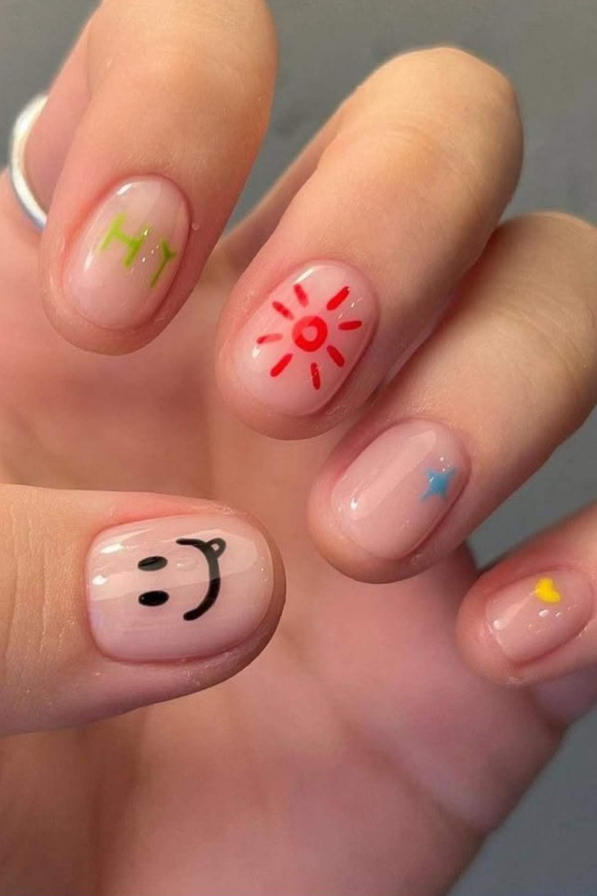 A hand with short, natural nails featuring colorful minimalist designs: "Hi", a sun, a smiley face, a blue star, and a yellow heart on a neutral base.