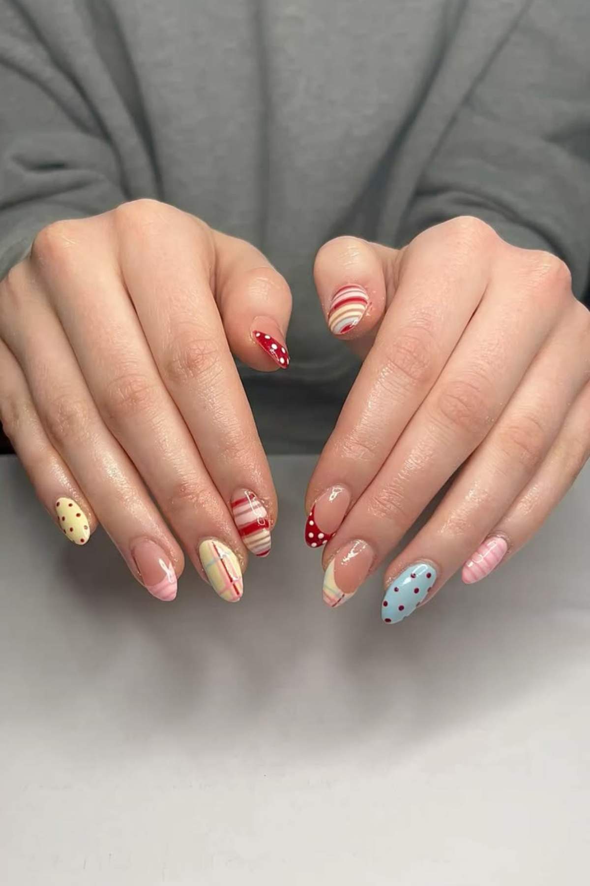 A person with manicured nails featuring various pastel-colored designs, including stripes, dots, and abstract patterns, holds their hands on a white surface.