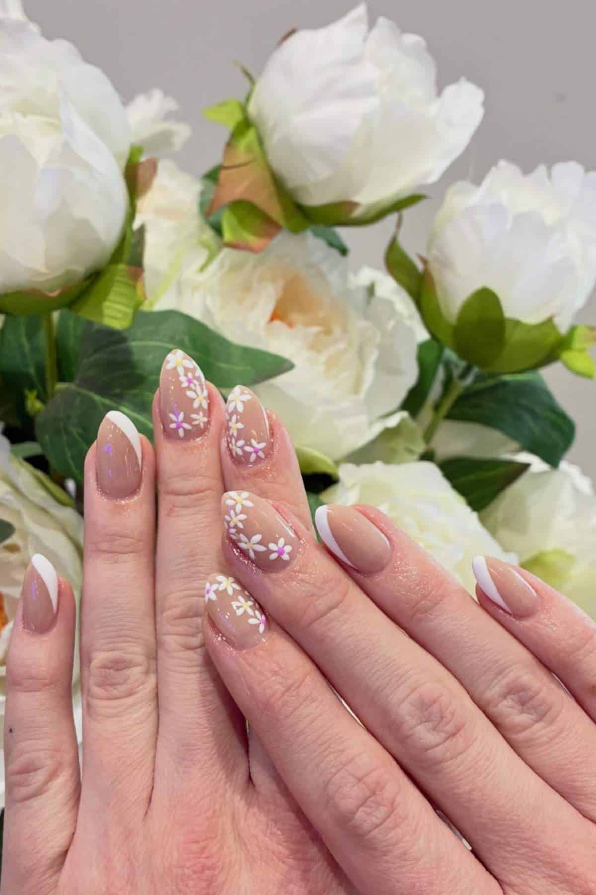 Hands with almond-shaped nails featuring a French tip and white and pink floral nail art, held in front of a bouquet of white flowers.